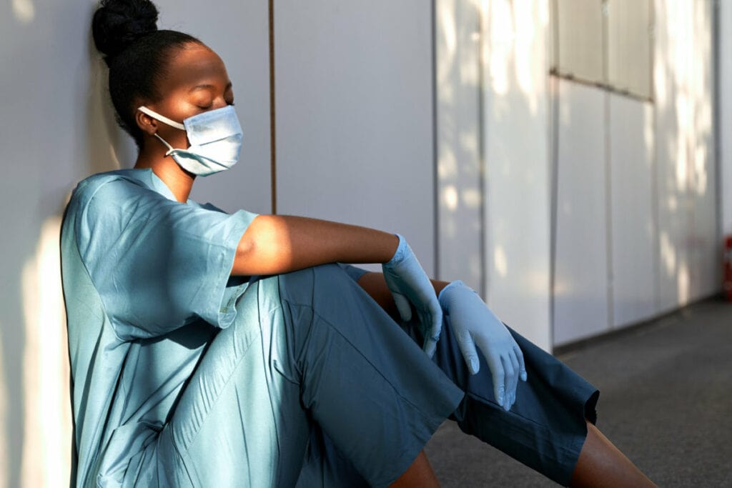 Tired scrub nurse wears face mask blue uniform gloves sits on hospital floor.